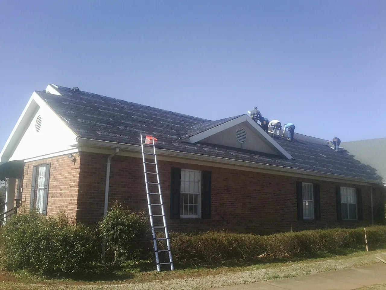 Crew tearing shingles off a brick ranch roof with a ladder set — Metal Roof Repair work by our crew in Jericho